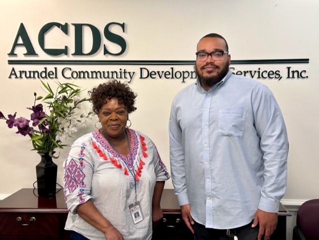 Two people stand and smile in front of a wall sign that reads