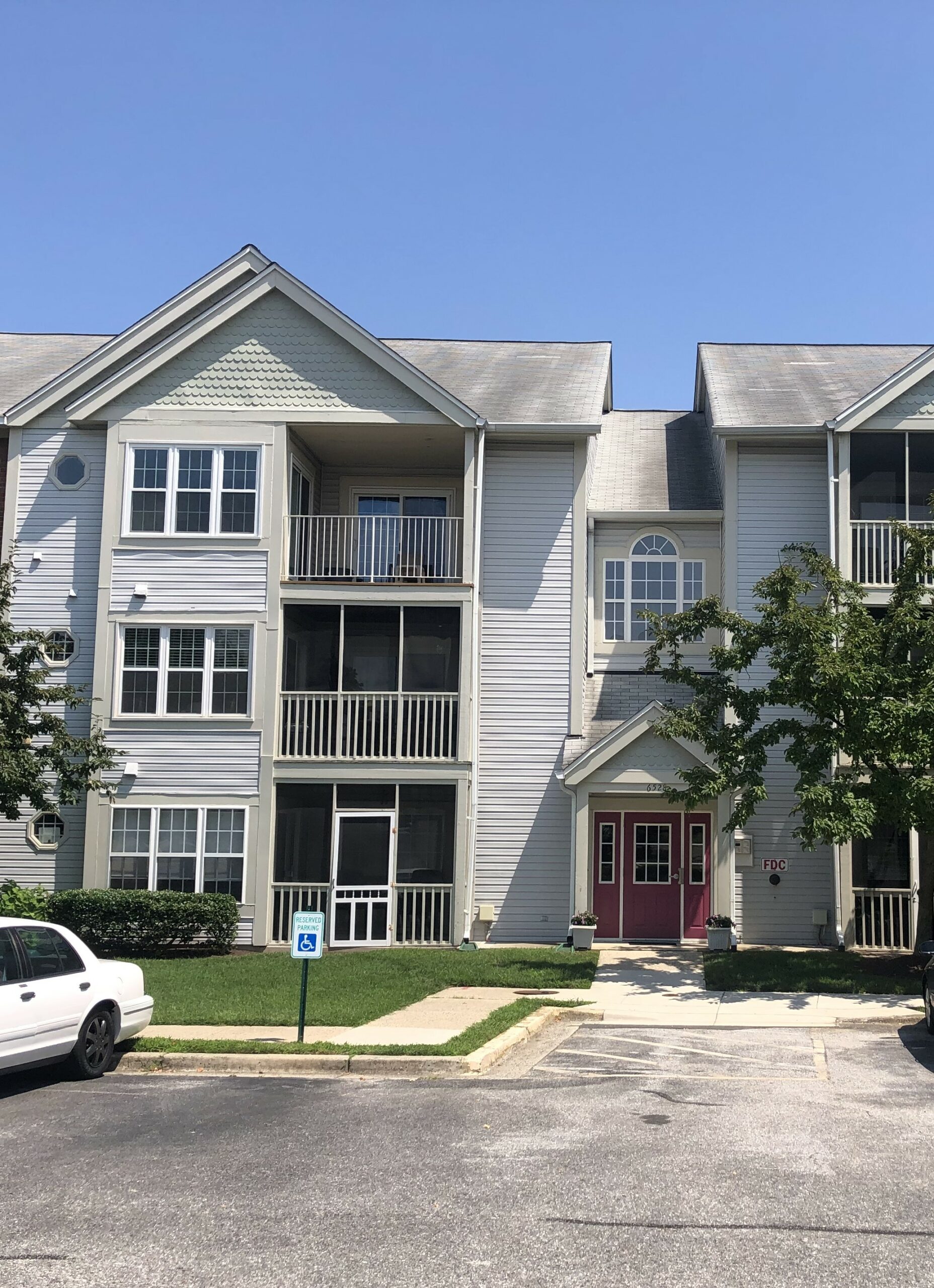 A three-story apartment building with light gray siding, balconies, red double front doors, and a small lawn. A white car is parked in front next to a handicap parking sign. Trees frame the entrance.