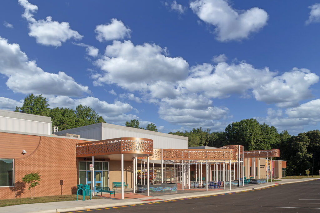 A modern school building with a brick facade, decorative orange canopy, large windows, blue benches, and trees in the background under a bright, partly cloudy sky.