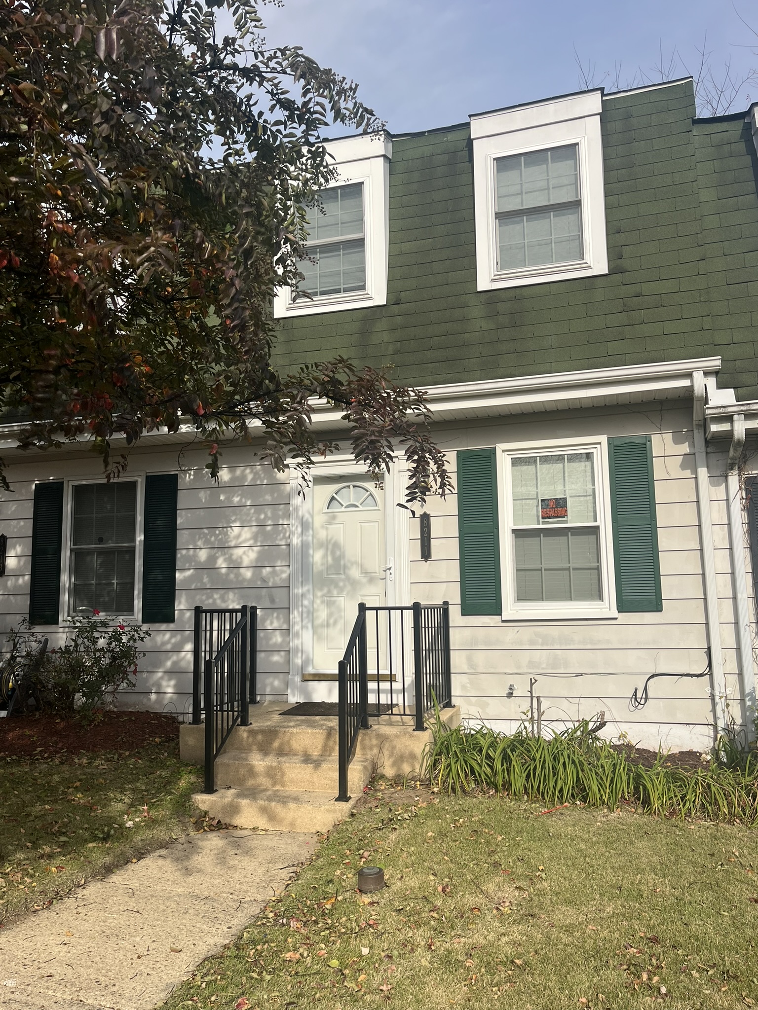 A two-story white house with green roof and shutters, black railings on steps leading to the front door, and a small yard with grass and some plants. A tree with red leaves partially frames the view.