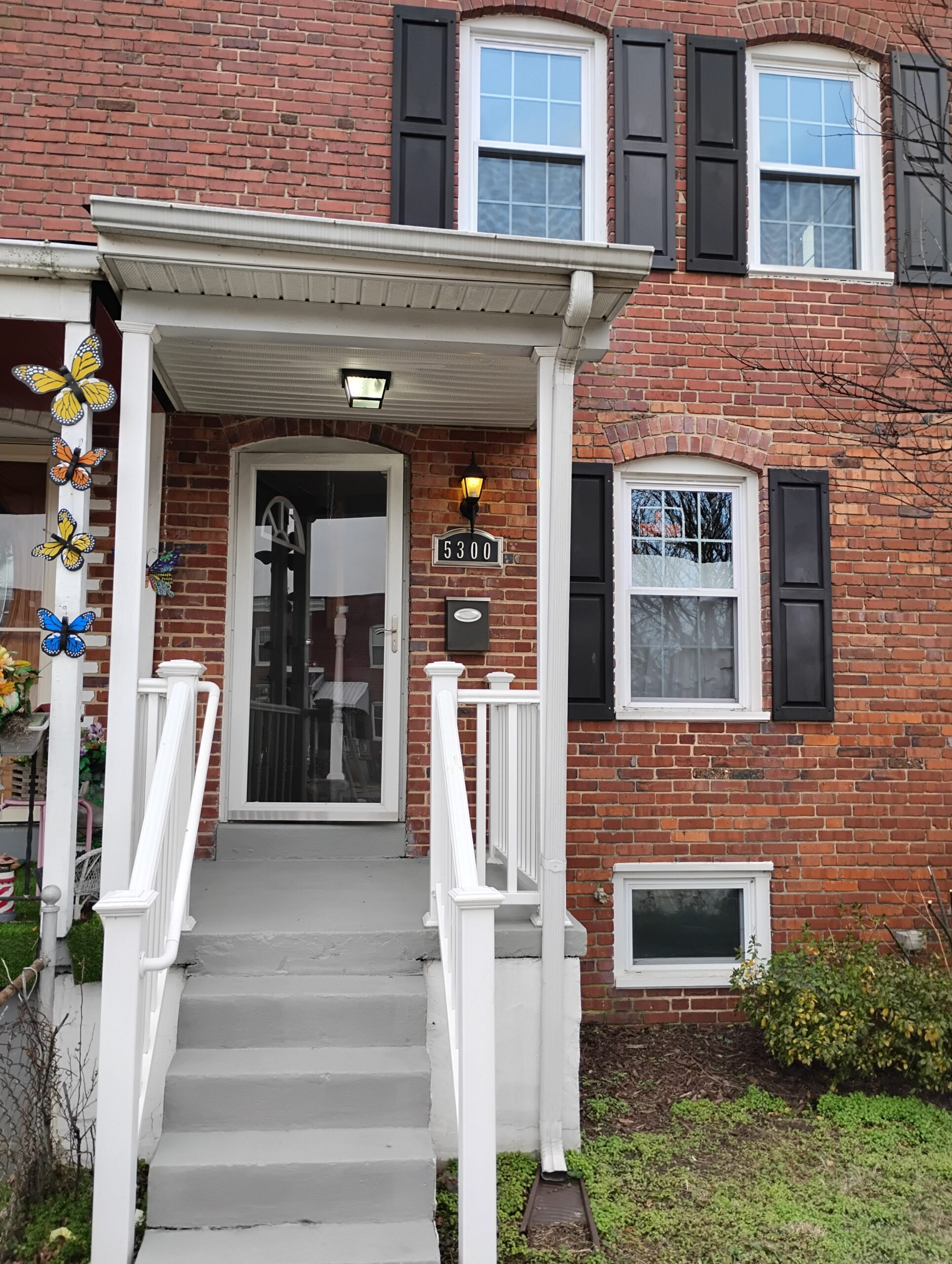 A brick townhouse with black shutters, white trim, and a covered front porch with steps and railings. House number 5300 is visible. Decorative butterflies are mounted on the porch and a window.