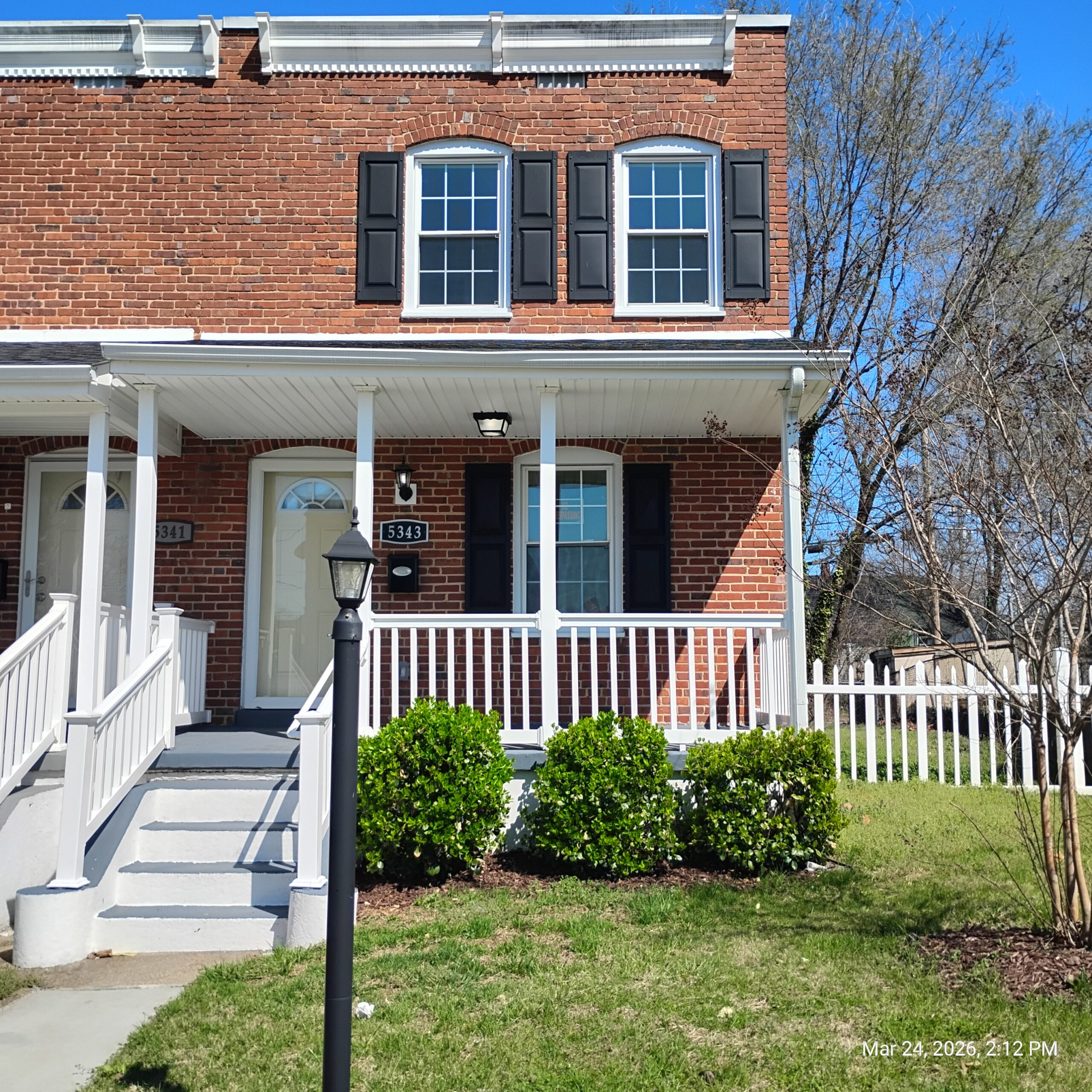A two-story brick house with black shutters, white trim, and a porch with white railings. Green bushes line the front, and a lamppost stands by the steps. Trees and a white fence are visible in the background.