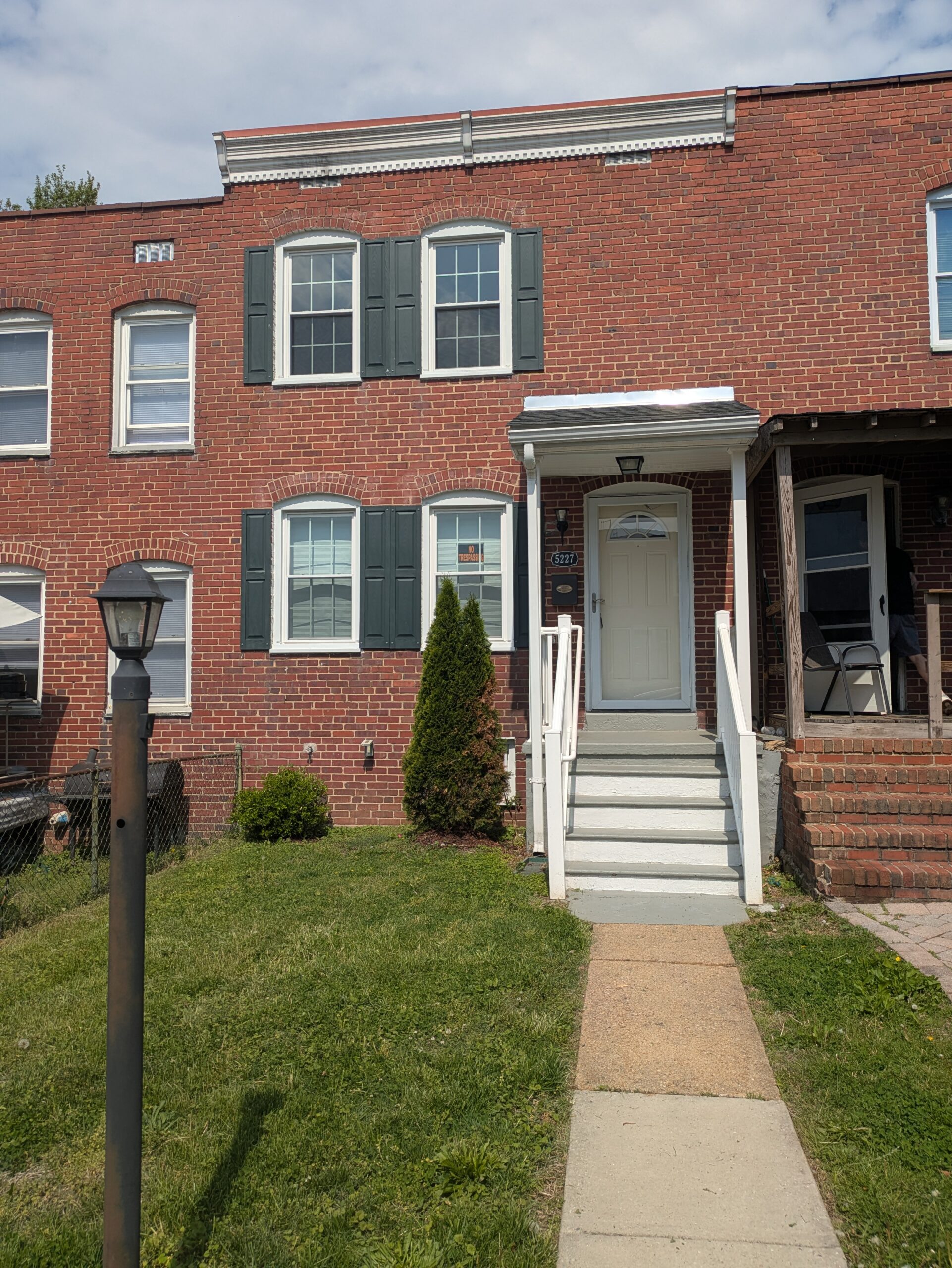 A red brick townhouse with green shutters, white trim, and a covered front porch. Steps lead up to a white front door. There is a grassy lawn, a small bush, and a lamp post in the yard under a partly cloudy sky.