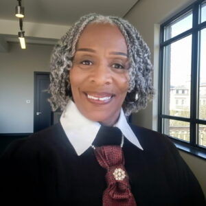 A smiling older woman with gray curly hair wears a white-collared shirt, black sweater, and a decorative red tie. She stands indoors near large windows overlooking a cityscape.