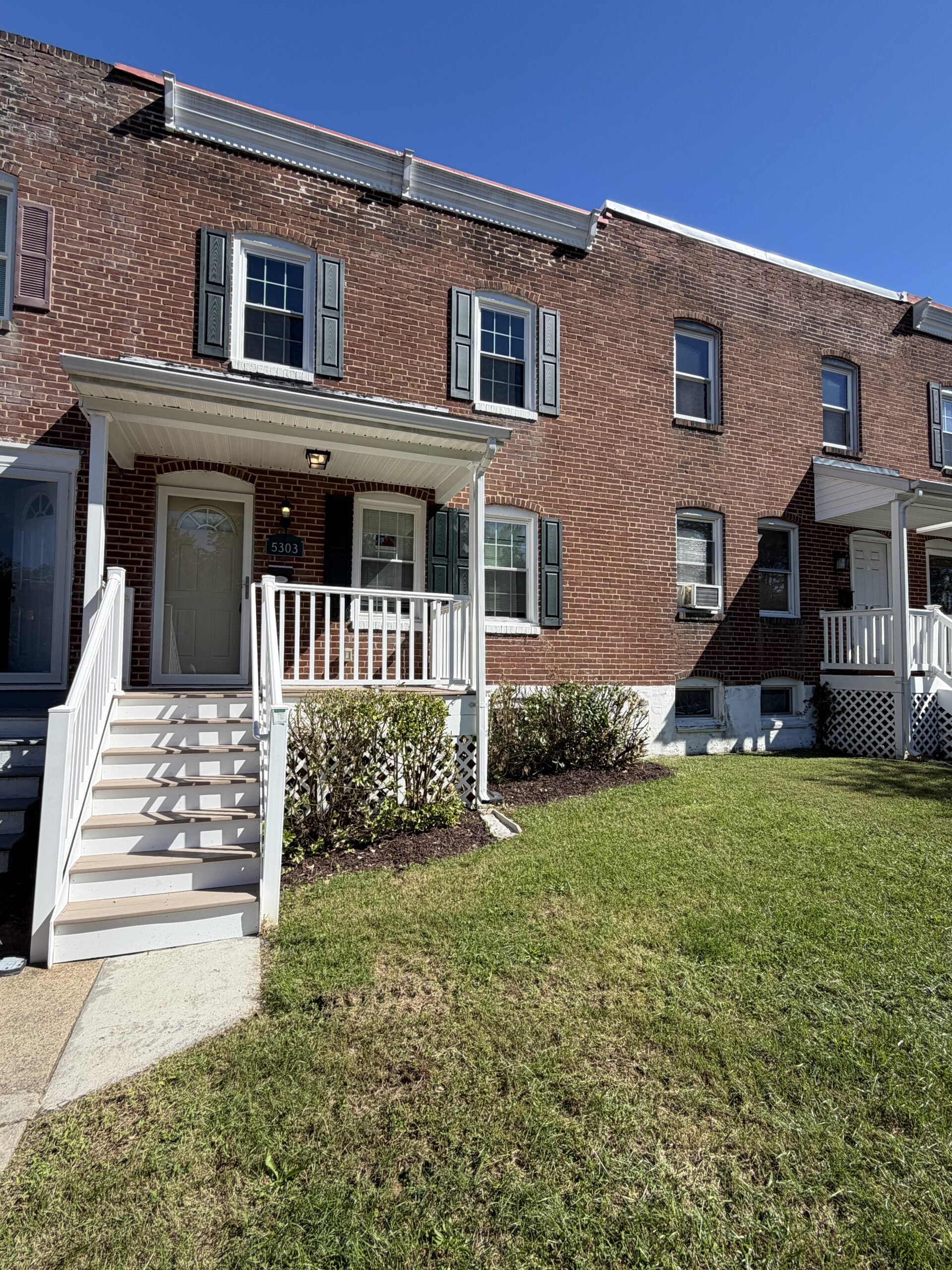 A two-story brick row house with a white front porch, stairs, and dark shutters under a clear blue sky.