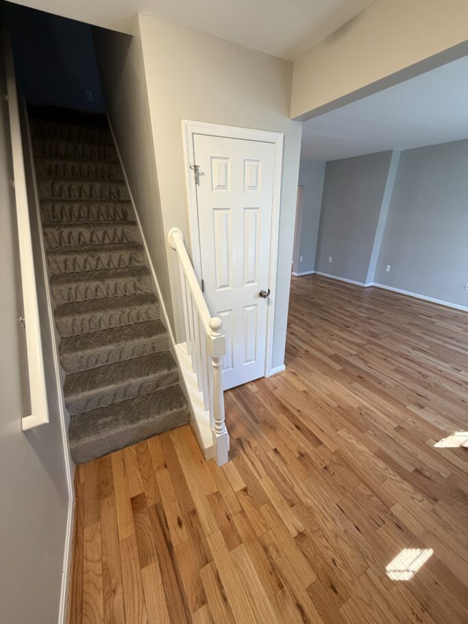 Stairs with grey carpet lead upward next to a white door and an empty room with hardwood floors.