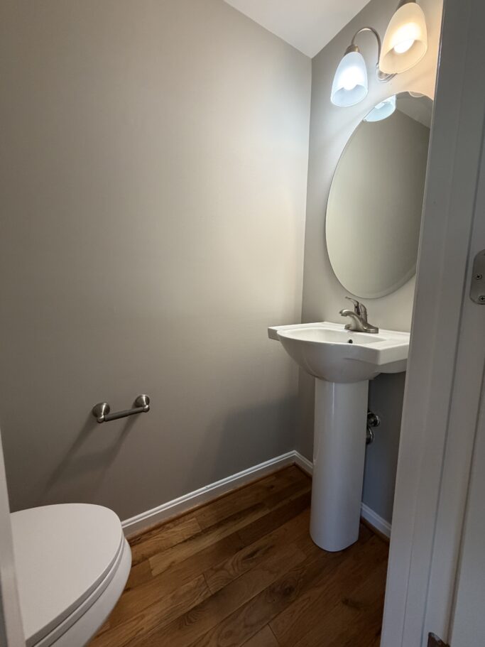 Small bathroom with a pedestal sink, oval mirror, wall-mounted light fixture, and a toilet.