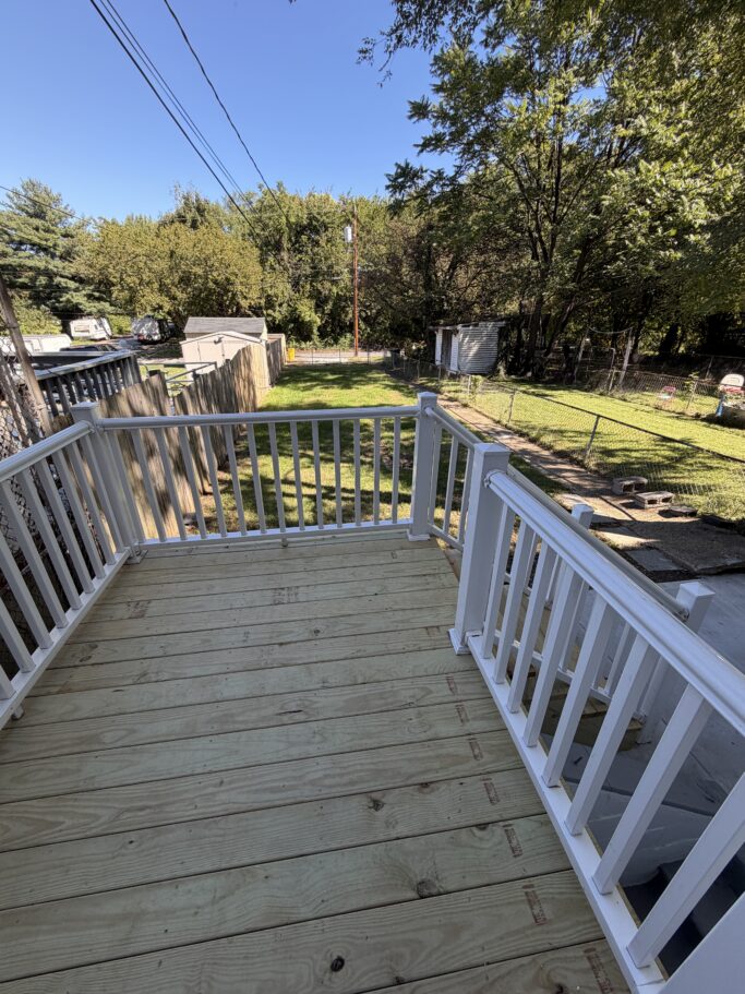 A newly constructed wooden deck with white railings overlooks a grassy backyard surrounded by trees and a wooden fence.