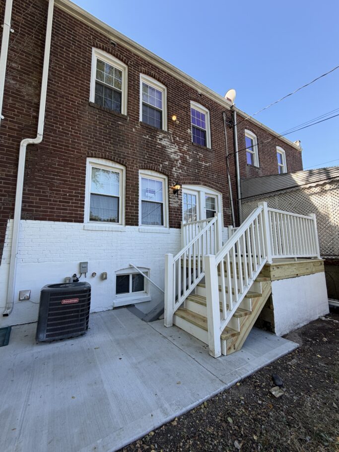 The rear exterior of a brick row house with white painted brick at the base, a concrete patio, a Goodman air conditioning unit, and a set of wooden stairs leading to a back door.
