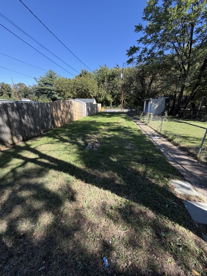 A narrow grassy backyard bordered by a wooden fence on the left and a chain-link fence with a concrete path on the right.