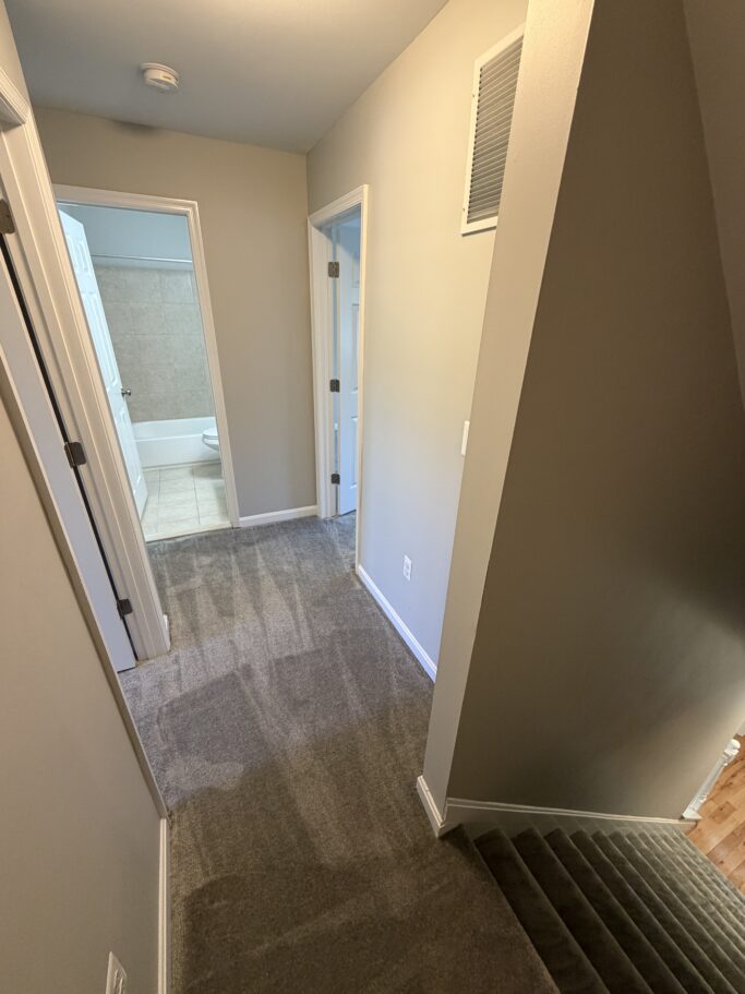 A view from the top of a staircase looking into a hallway with two open doors, one leading to a bathroom.