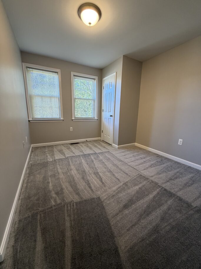 An empty room with light gray walls, gray patterned carpet, two windows with blinds, and a closed white door.
