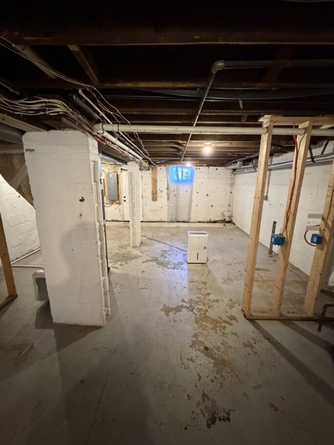 An unfinished basement with concrete floors, white-painted foundation walls, exposed ceiling joists, and a dehumidifier sitting in the center of the room.