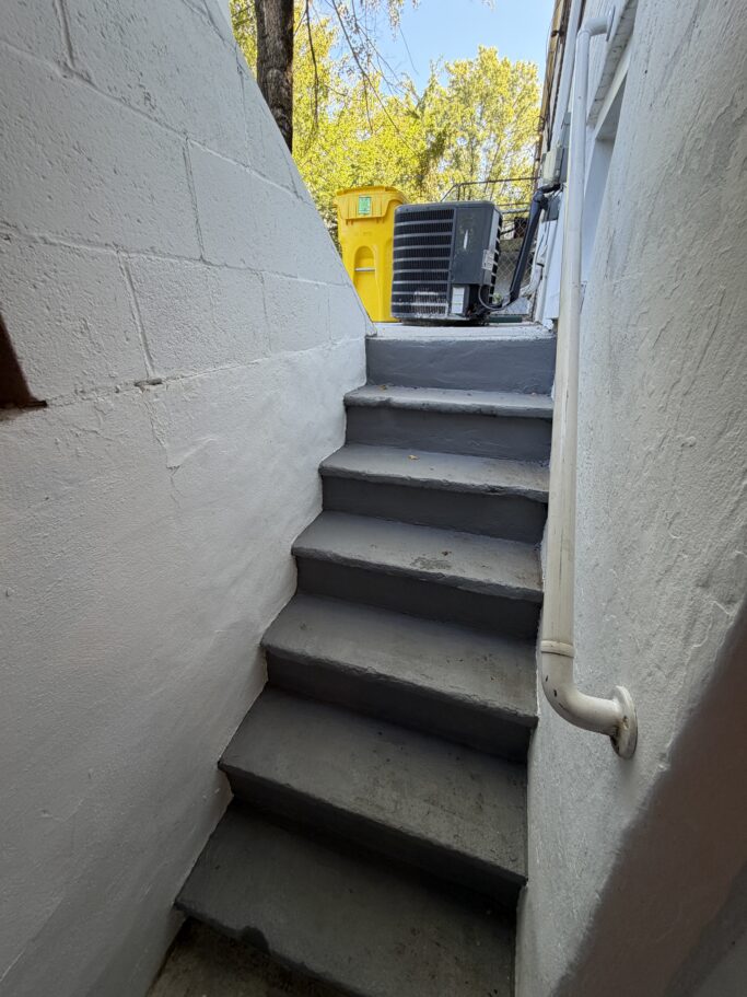 Concrete stairs lead upward between white walls toward an outdoor air conditioning unit and a yellow trash bin.
