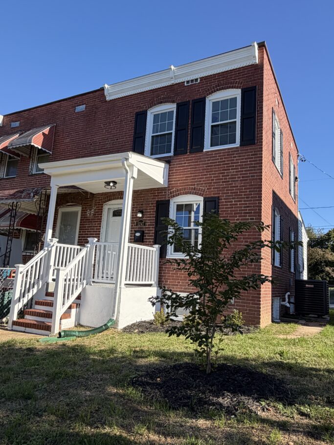 Two-story red brick row house with a white front porch, black shutters, and a small tree in the front yard.