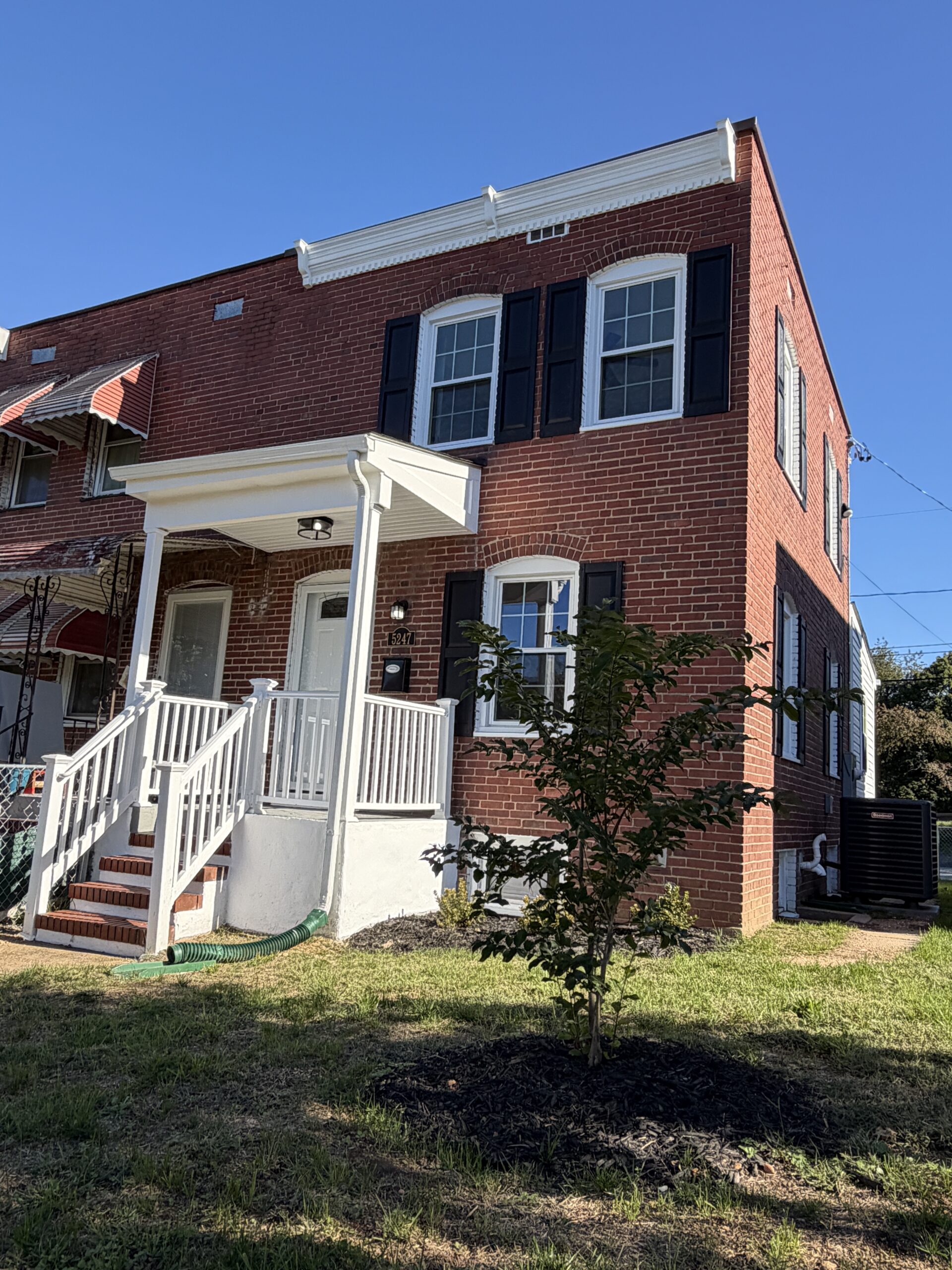 Two-story red brick row house with a white front porch, black shutters, and a small tree in the front yard.