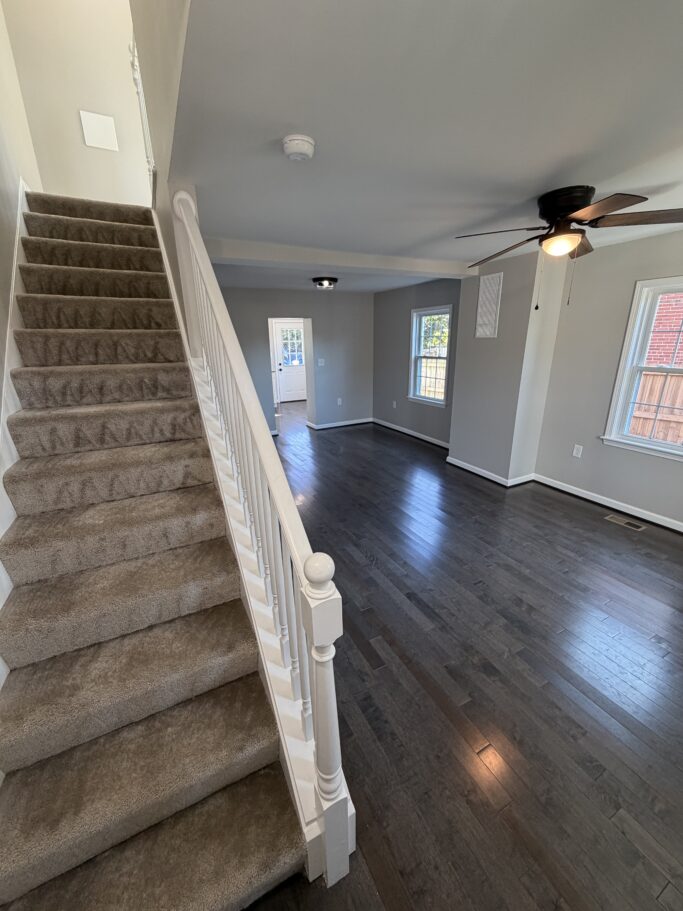 A carpeted staircase with a white railing leading down to an open living area with dark hardwood floors and windows.
