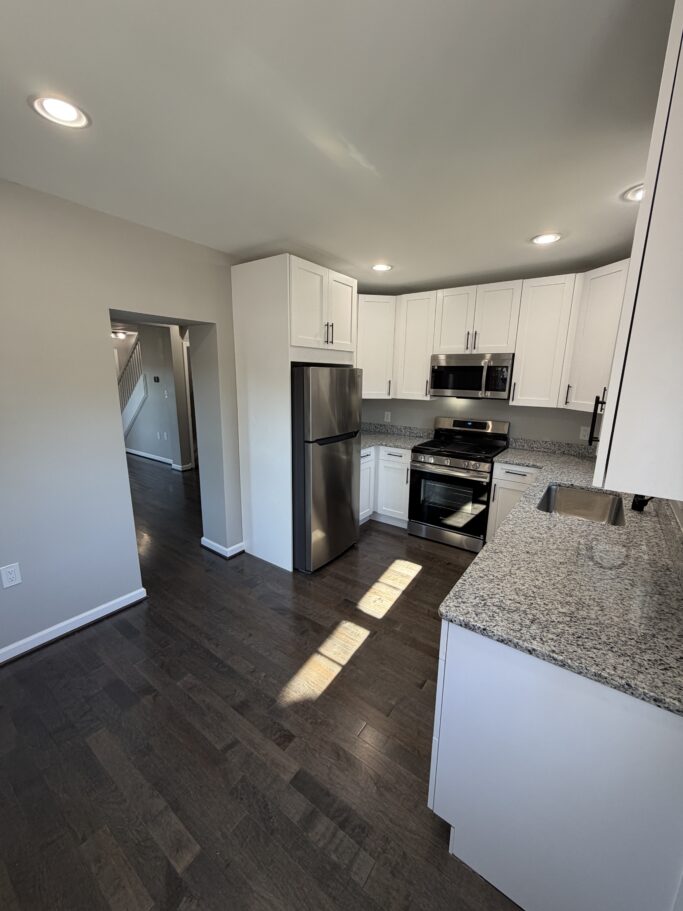 A kitchen with white cabinets, granite countertops, stainless steel appliances, and dark hardwood floors, with a doorway leading to a staircase in the background.
