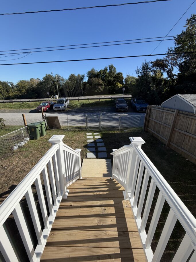 A wooden staircase with white railings leads down from a deck to a backyard with a path of square pavers and a metal fence bordering a parking area.