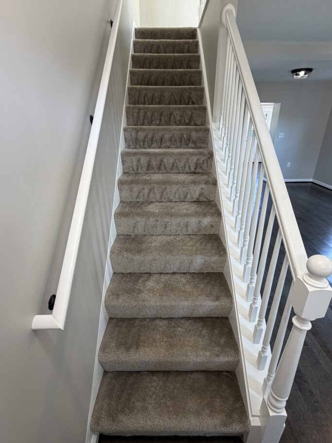 Straight staircase with beige carpet, a white handrail on the left, and white balusters on the right.