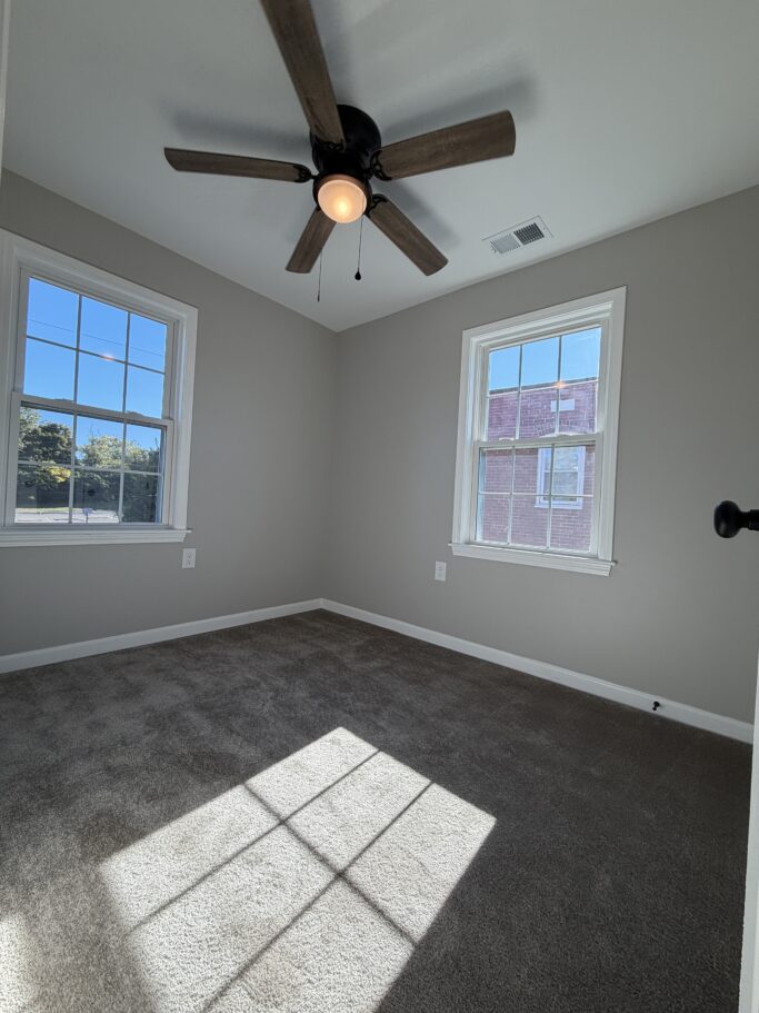 An empty bedroom with gray walls, carpeted floors, two windows, and a ceiling fan.