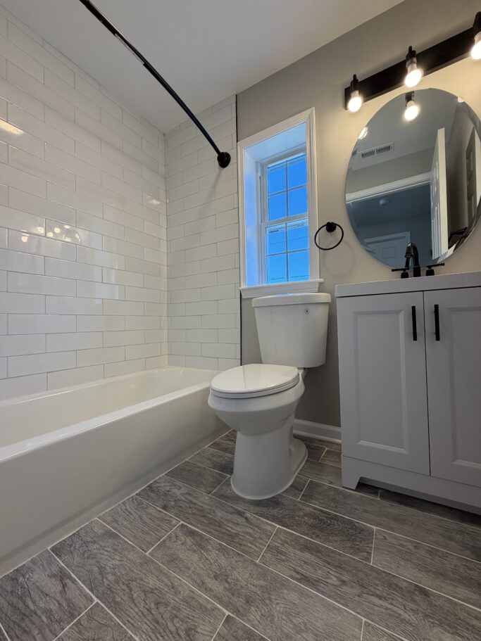A bathroom featuring a white subway-tile shower, a toilet, a gray vanity with a round mirror, and wood-look tile flooring.