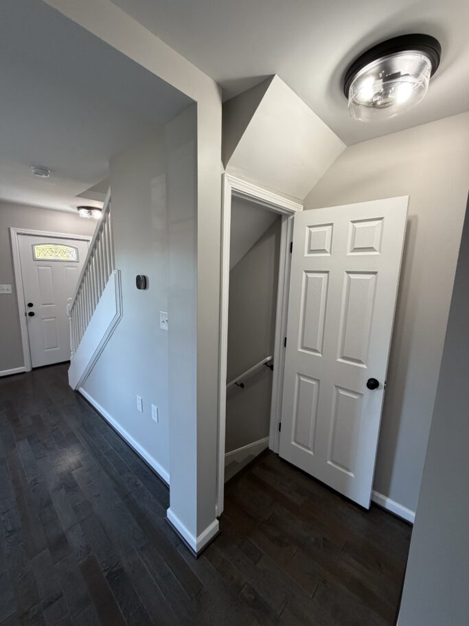A hallway with dark wood floors showing a staircase and an open door to a basement or storage area.