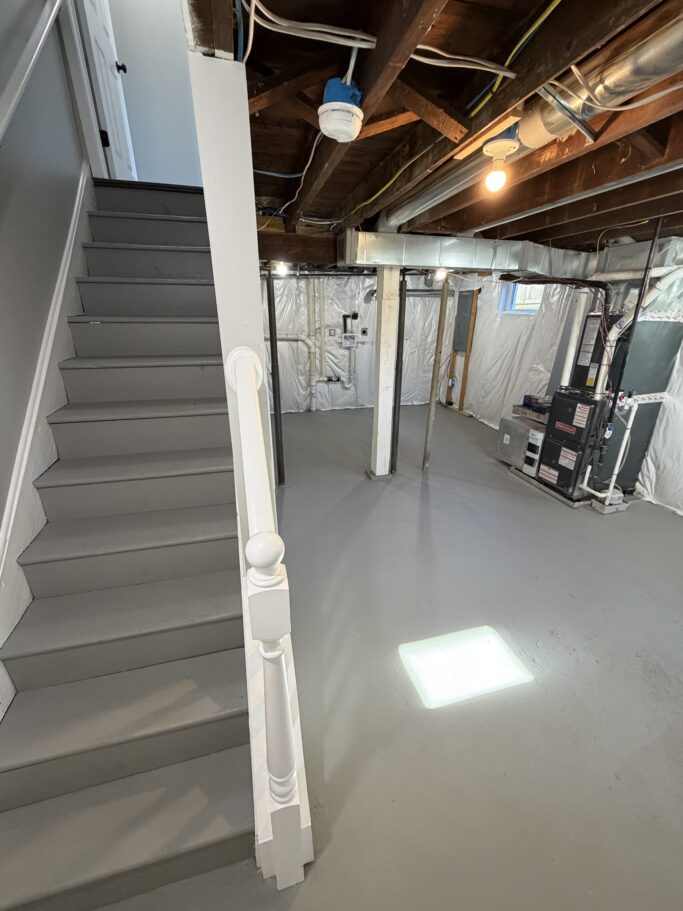 Basement interior with gray painted stairs leading up, a painted gray floor, and an exposed ceiling with HVAC equipment.